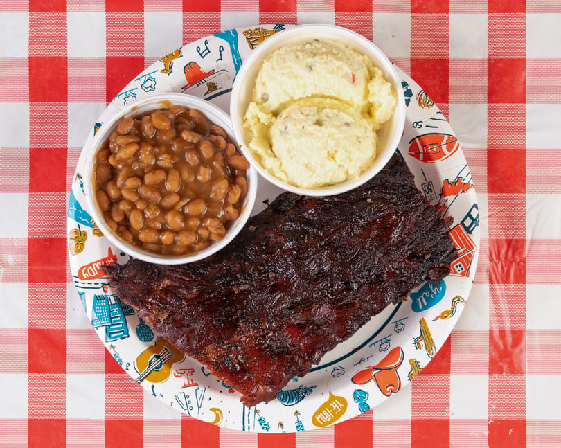 A rack of BBQ ribs, baked beans, and potato salad on a paper plate over a red and white checkered tablecloth.