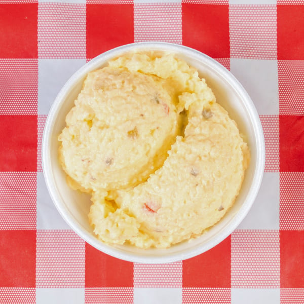Creamy potato salad with red and green peppers in a white bowl on a red and white checkered tablecloth.
