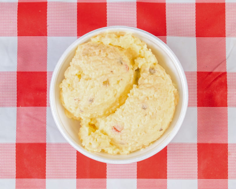 Creamy potato salad with red and green peppers in a white bowl on a red and white checkered tablecloth.