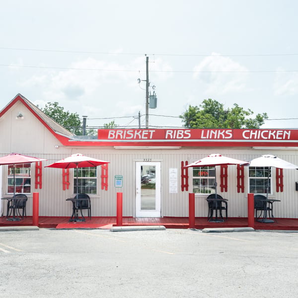 Exterior of a small, light-colored building with red trim, outdoor seating, and a sign for "Brisket Ribs Links Chicken."