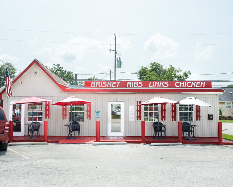 Exterior of a small, light-colored building with red trim, outdoor seating, and a sign for "Brisket Ribs Links Chicken."