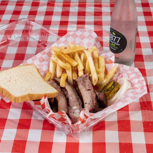 BBQ ribs, fries, white bread, pickles, and onions in a plastic container on a red and white checkered tablecloth.