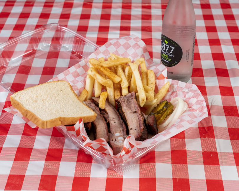 BBQ ribs, fries, white bread, pickles, and onions in a plastic container on a red and white checkered tablecloth.