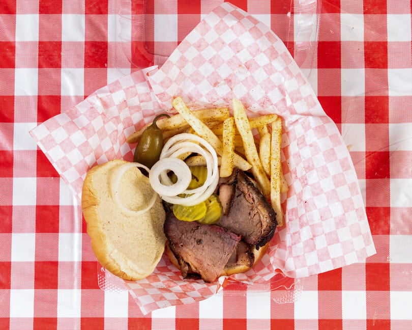 Top-down view of a brisket sandwich with fries, onions, pickles, and a jalapeño on a red and white checkered tablecloth.