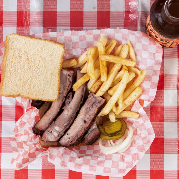 Rib plate with fries, white bread, pickles, and onions on a red and white checkered tablecloth.