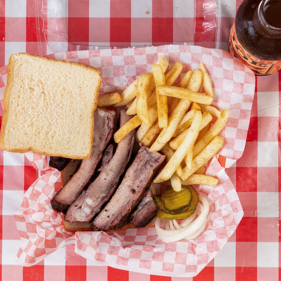 Rib plate with fries, white bread, pickles, and onions on a red and white checkered tablecloth.