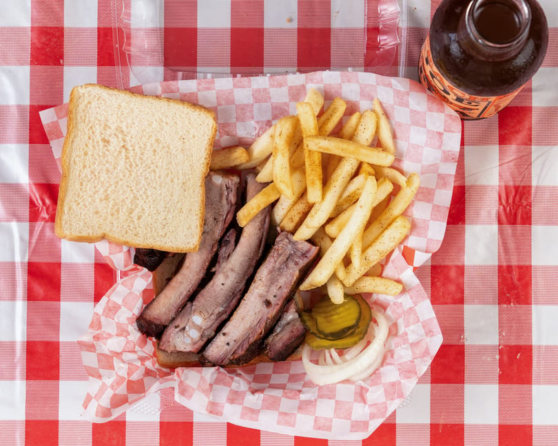 Rib plate with fries, white bread, pickles, and onions on a red and white checkered tablecloth.