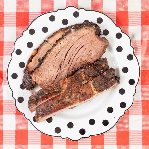 Slices of smoked brisket and ribs on a polka-dotted plate over a red and white checkered tablecloth.