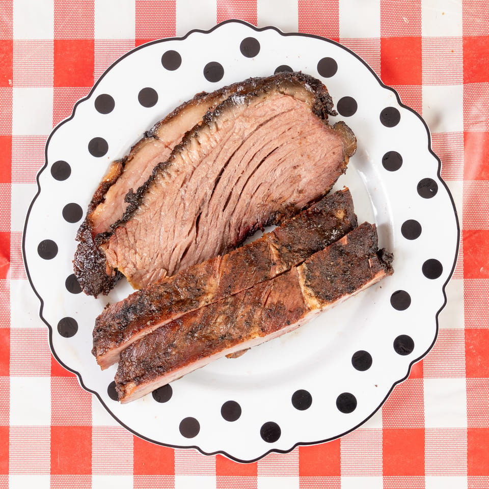 Slices of smoked brisket and ribs on a polka-dotted plate over a red and white checkered tablecloth.