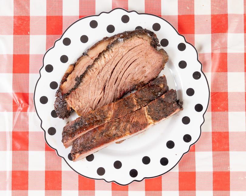 Slices of smoked brisket and ribs on a polka-dotted plate over a red and white checkered tablecloth.