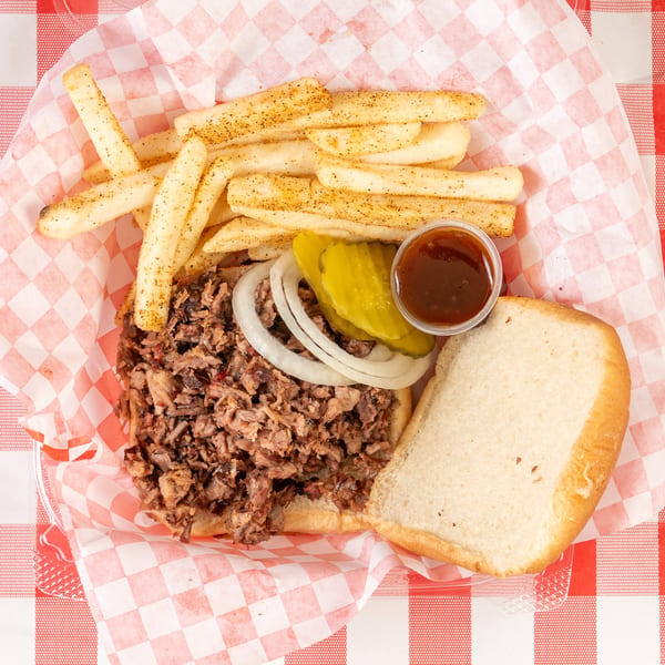 Pulled pork sandwich with onions, pickles, BBQ sauce, and seasoned fries in a red checkered basket.