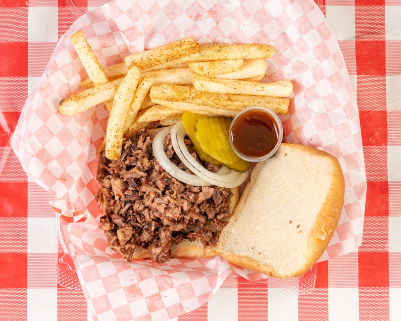 Pulled pork sandwich with onions, pickles, BBQ sauce, and seasoned fries in a red checkered basket.