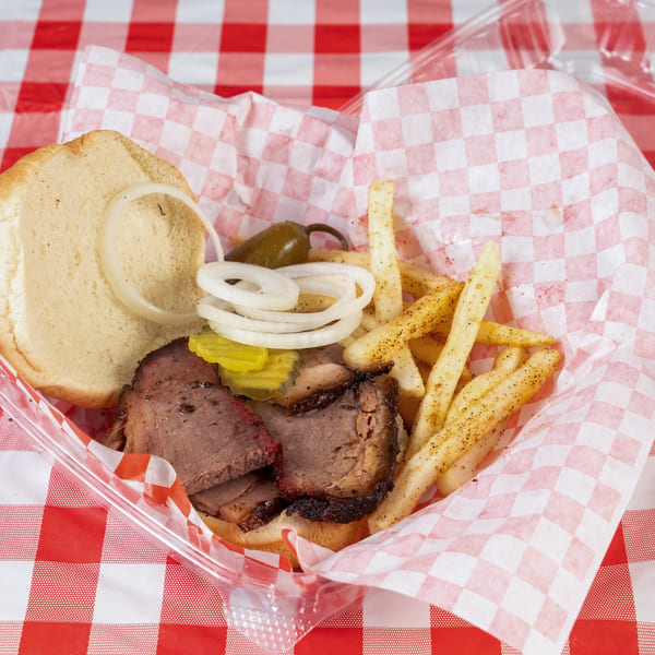 Brisket sandwich with onions, pickles, and a jalapeño, served with seasoned fries in a plastic container.