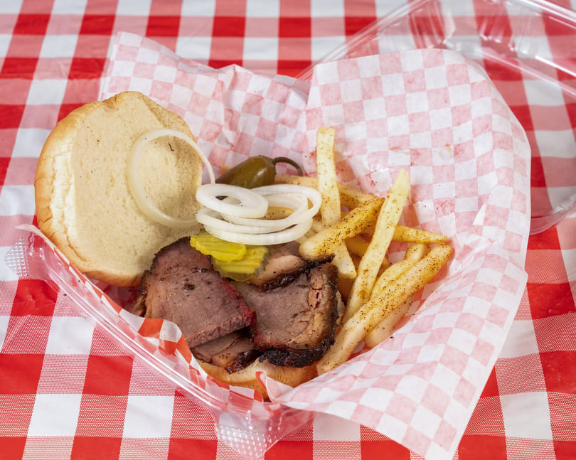 Brisket sandwich with onions, pickles, and a jalapeño, served with seasoned fries in a plastic container.