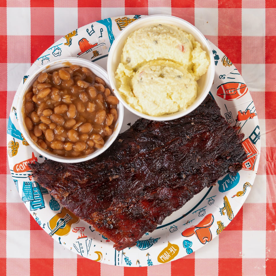 A rack of BBQ ribs, baked beans, and potato salad on a paper plate over a red and white checkered tablecloth.