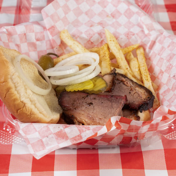 Brisket sandwich with onions, pickles, and fries in a red and white checkered basket.