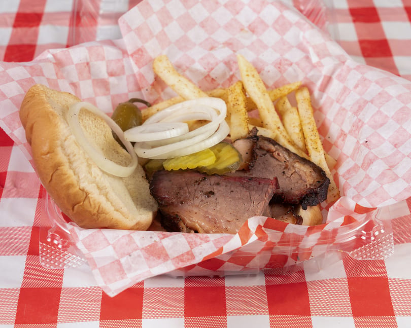 Brisket sandwich with onions, pickles, and fries in a red and white checkered basket.