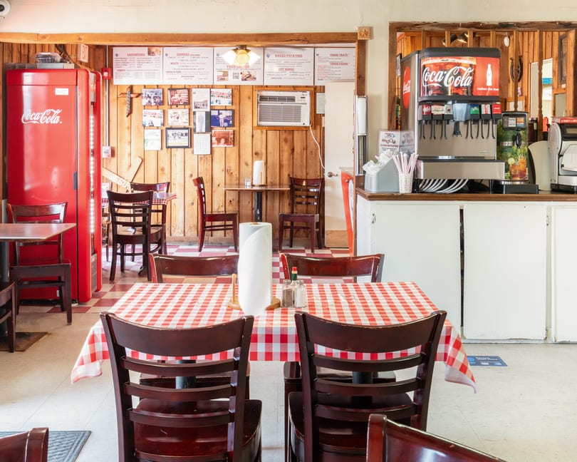 Interior of a casual restaurant with wooden walls, red and white checkered tablecloths, and a Coca-Cola machine.