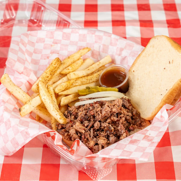 Pulled pork sandwich with fries, pickles, onions, and BBQ sauce in a plastic container on a red and white checkered tablecloth.