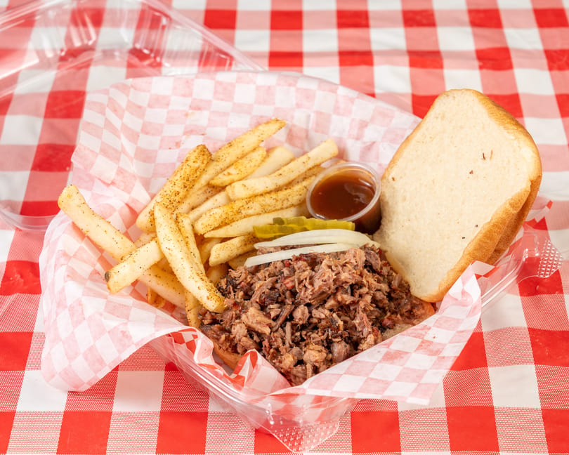 Pulled pork sandwich with fries, pickles, onions, and BBQ sauce in a plastic container on a red and white checkered tablecloth.