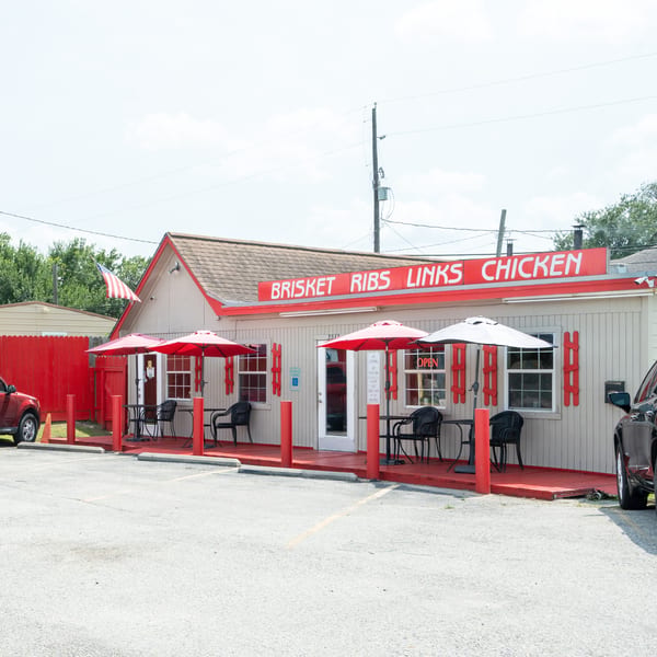 Exterior of a small, light-colored restaurant with a red roof, deck, and umbrellas, with cars parked.