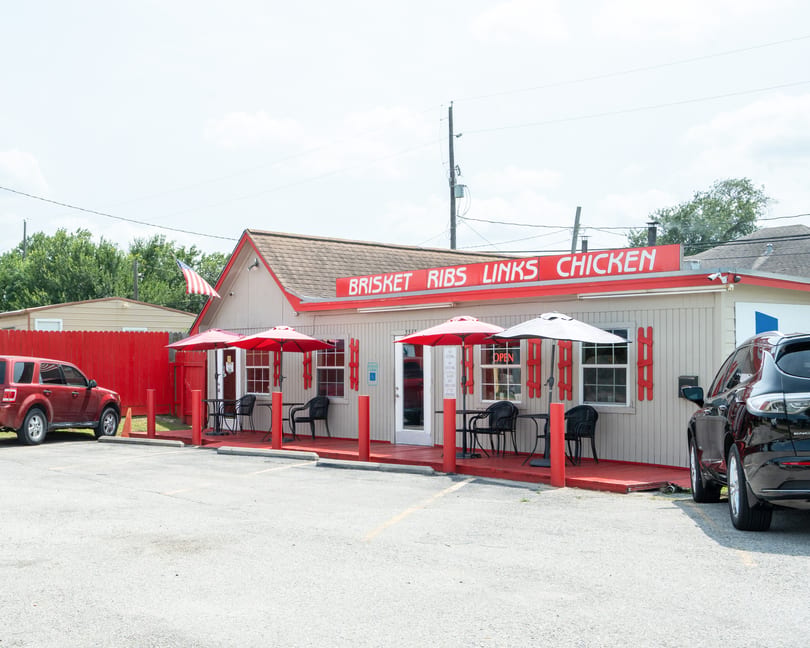 Exterior of a small, light-colored restaurant with a red roof, deck, and umbrellas, with cars parked.